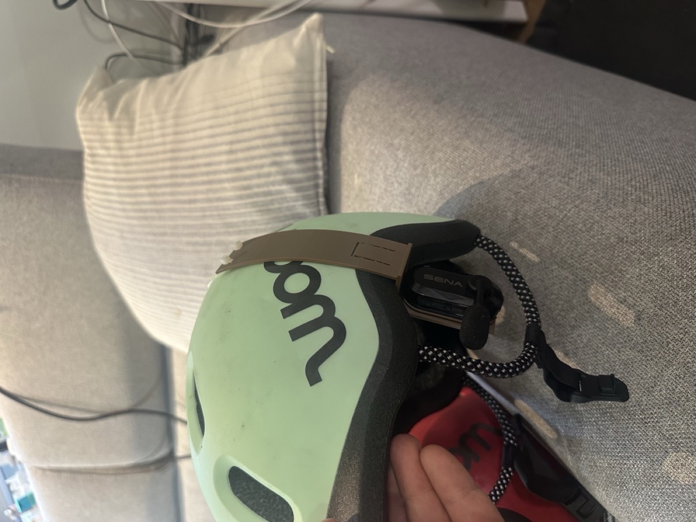 The beige printed clip hooked over the rim of a mint woom helmet, the black Sena unit hanging under the rim by the ear
